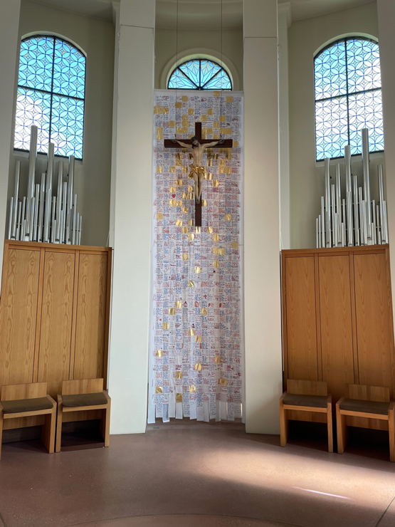 Das Hungertuch der Fastenzeit ist nun ein Ostertuch. Goldene Flächen durchziehen das Tuch hinter dem Kreuz. 